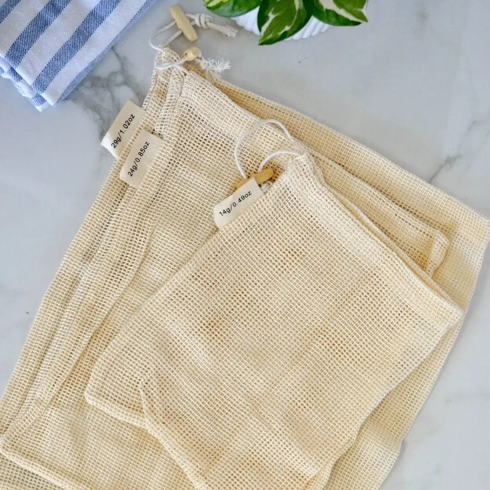 Set of beige mesh bags on a marble surface with a plant and towel in the background.