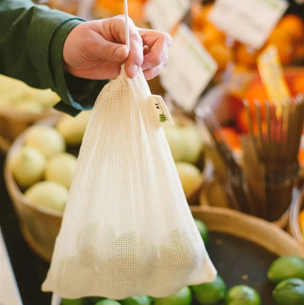 Hand holding a reusable mesh bag over a produce section with fruits and vegetables.