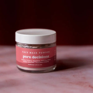 Jar of 'Pore Decisions' face mask powder on a pink marble surface with a dark background