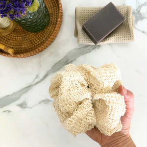 Hand holding a loofah sponge with soap bar and flowers on marble surface