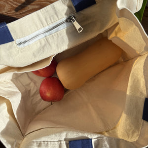 Beige reusable tote bag with blue handles containing multiple pockets and a pocket with a zipper, filled with 2 apples and a butternut squash.