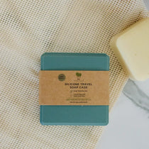 A teal silicone travel soap case with a kraft paper band label, placed on a textured cream mesh fabric next to a bar of soap.