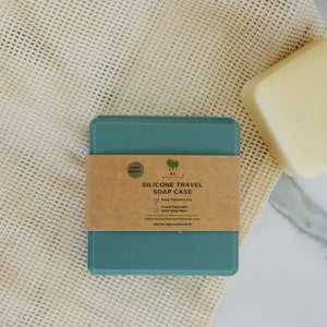 A teal silicone travel soap case with a kraft paper band label, placed on a textured cream mesh fabric next to a bar of soap.