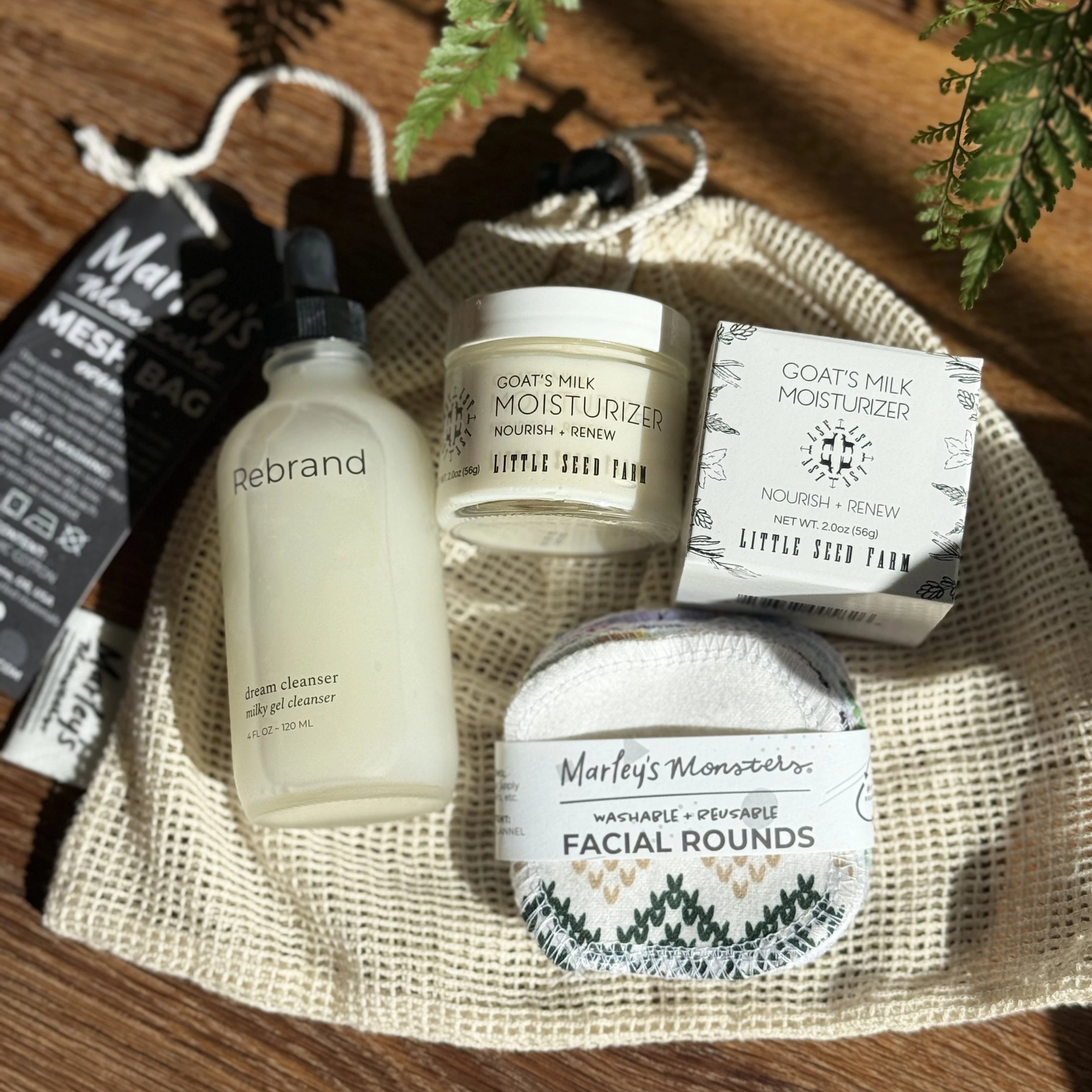 Facial cleanser glass bottle, reusable facial rounds and goat milk moisturizer laying on a cotton mesh bag on a wooden surface
