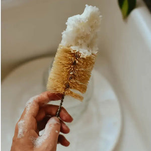Hand holding a natural bristle brush with soap suds over a sink.