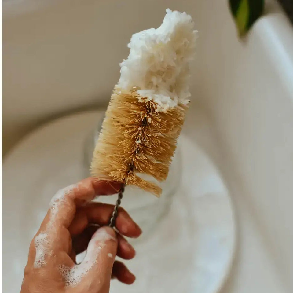 Hand holding a natural bristle brush with soap suds over a sink.