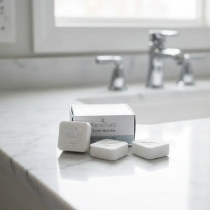 Three white MamaSuds Toilet Bombs are arranged in front of their white and gray product box on a white marble bathroom countertop, with a blurred sink and faucet in the background.