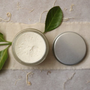 Jar of organic shea butter with a metal lid and green leaves on a textured fabric background