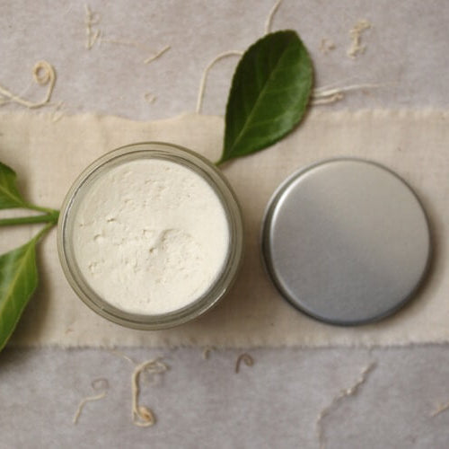 Jar of organic shea butter with a metal lid and green leaves on a textured fabric background