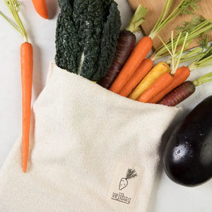 A beige canvas reusable produce bag with a sewn-on 'vejibag' logo, filled with fresh vegetables including carrots, kale, and eggplant, arranged on a white marble surface with a wooden cutting board.