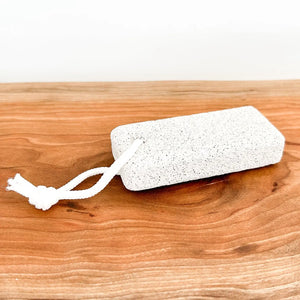 A rectangular white pumice stome with a white hanging cord, resting on a wooden surface.