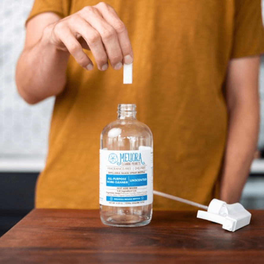 Person adding a tablet to an open and empty spray bottle labeled 'Meliora all-purpose cleaner' on a wooden surface