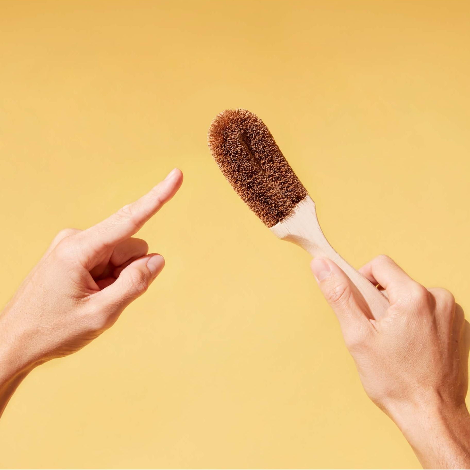 A person's hands holding and pointing to a cleaning brush with coconut husk bristles and a wooden handle against a solid yellow background.