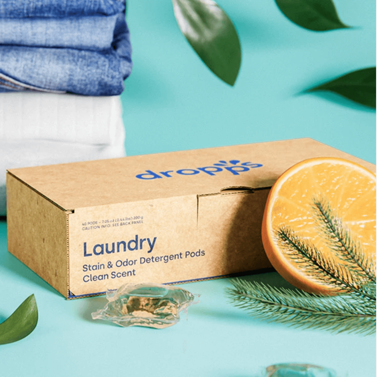 A box of Dropps Laundry Stain & Odor Detergent Pods with a laundry load in the background. The box is labeled 'Clean Breeze' scent and the visual suggests a fresh, clean laundry scent.
