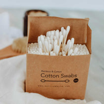 A compostable cardboard box filled with bamboo and cotton earbuds, placed on a white surface with a brush and towel in the background.