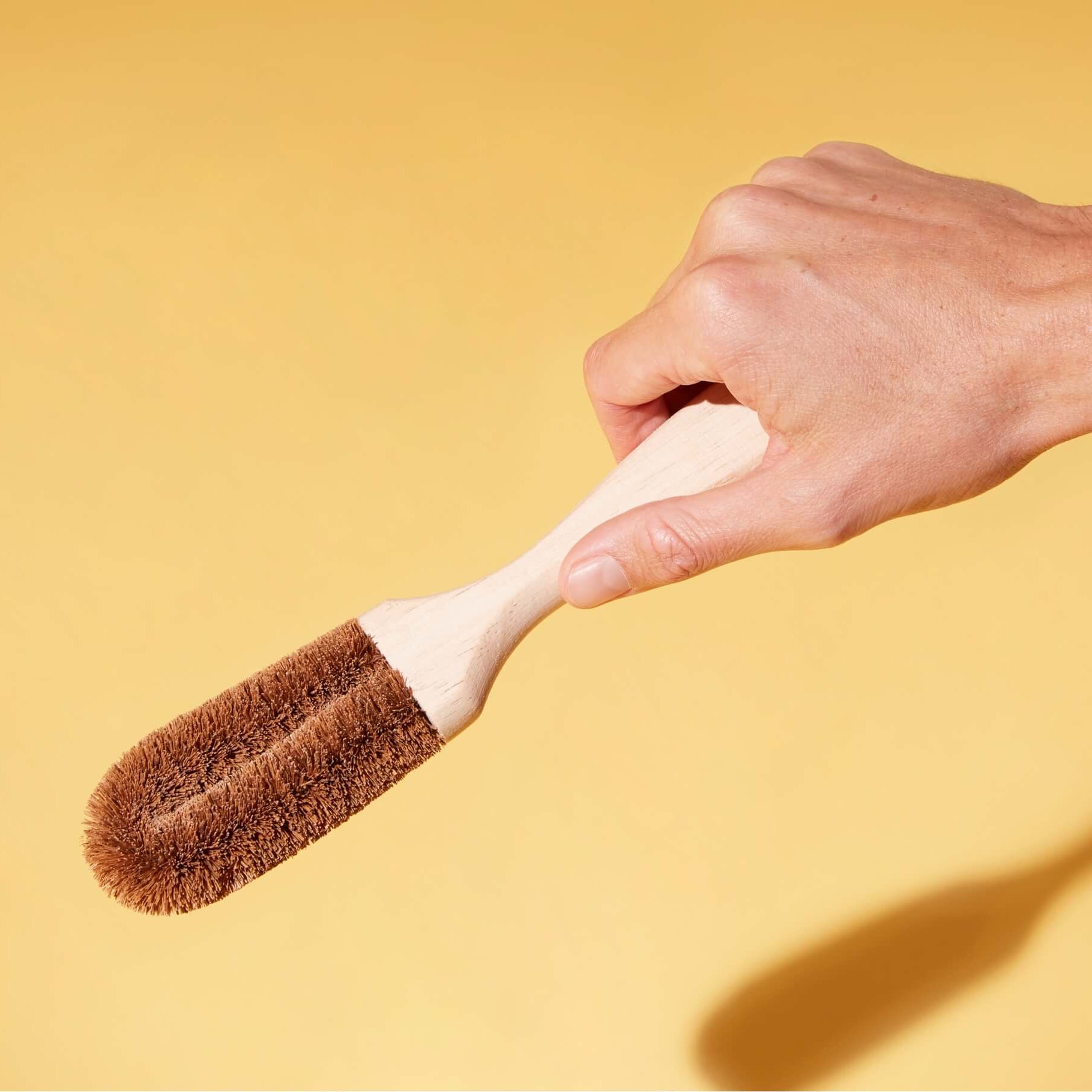 A hand holding a cleaning brush with coconut husk bristles and a wooden handle against a solid yellow background.