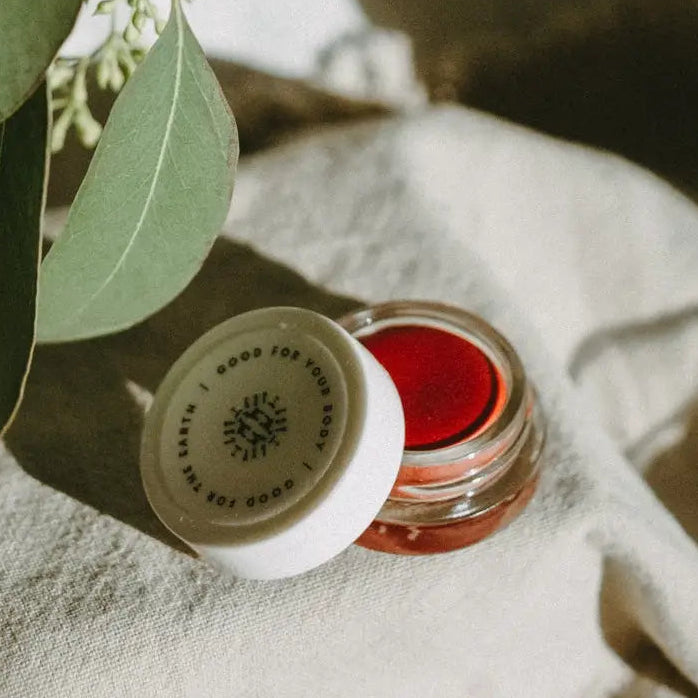A glass jar with a white lid containing red lip tint, placed on a soft, light environment, possibly indicating a cosmetic product setting.
