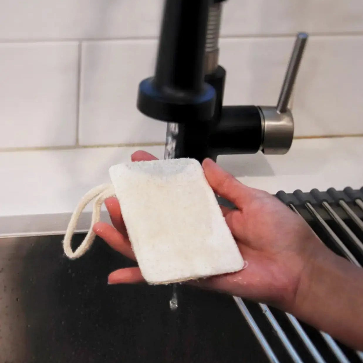 A hand holding a white loofah sponge under a kitchen tap, with a sponge mop in the background.