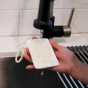 A hand holding a white loofah sponge under a kitchen tap, with a sponge mop in the background.