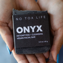 A hand holding a black charcoal cleansing bar with 'ONYX Detoxifying + Cleansing Vegan Facial Bar' printed on it.