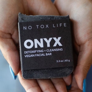 A hand holding a black charcoal cleansing bar with 'ONYX Detoxifying + Cleansing Vegan Facial Bar' printed on it.