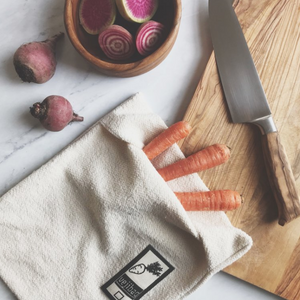 A beige Vejibag made of organic cotton, with carrots and a knife on a wooden board, alongside a small bowl of sliced radishes.