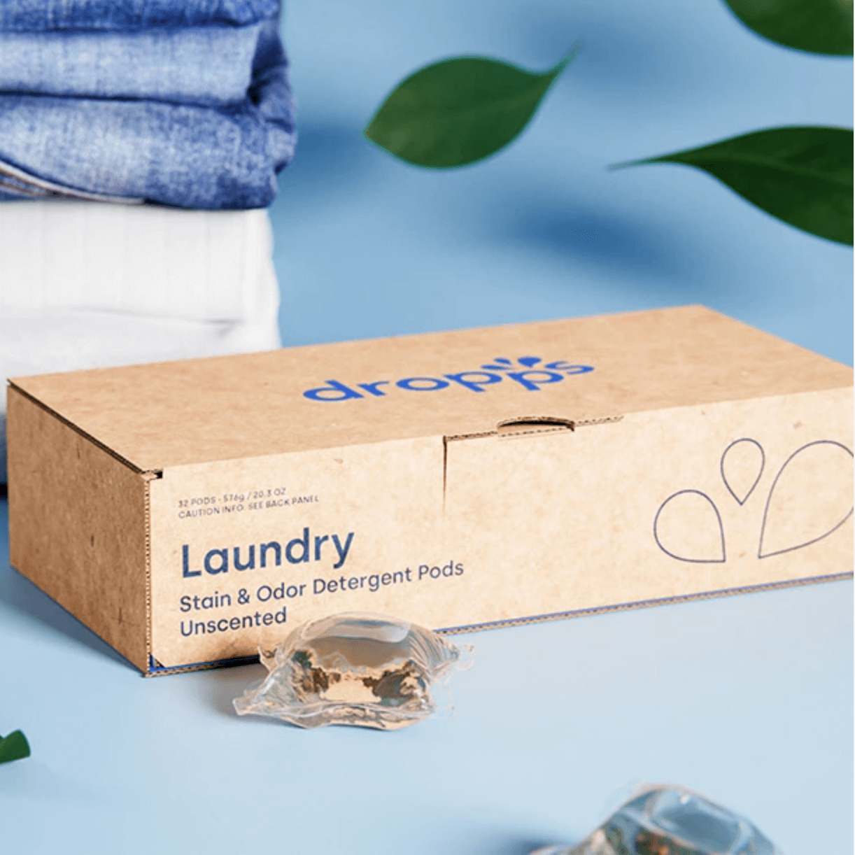 A box of Dropps Laundry Detergent Pods with a single pod visible, alongside a stack of white and blue clothes, and decorative elements like leaves and a slice of orange.