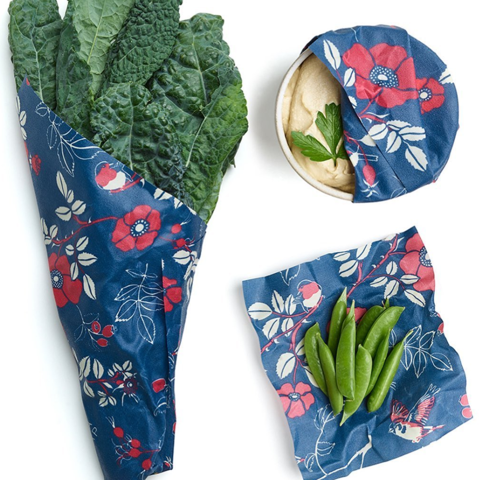 Assorted sizes of Bee's Wrap food storage wraps displayed on a kitchen counter with one small, one medium, and one large wrap shown partially covering food items. The wraps are green with a pattern of bees and pollen.