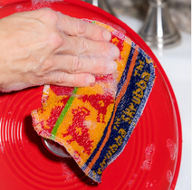 A person cleaning a red plate with a colorful, patterned scrub cloth.