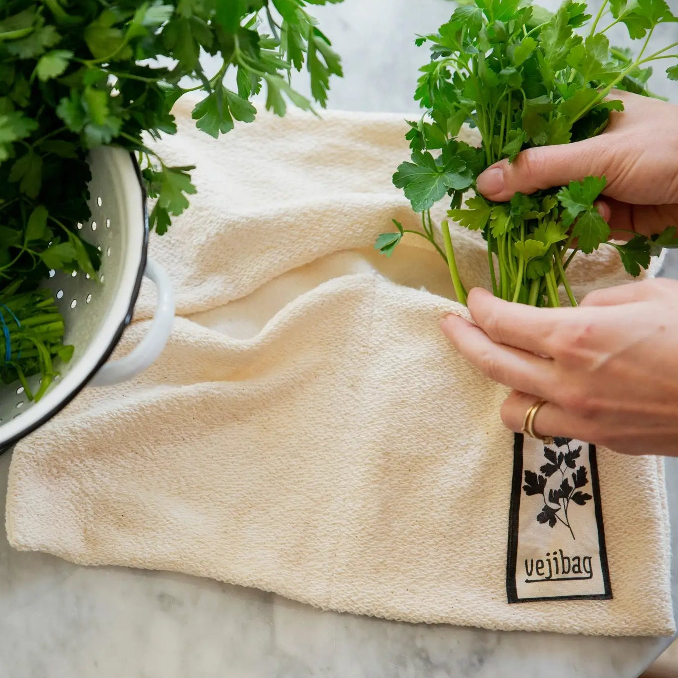A beige terrycloth kitchen towel with a black and white 'vejibag' brand tag, shown with hands holding fresh green parsley over a white marble countertop next to a metal colander.